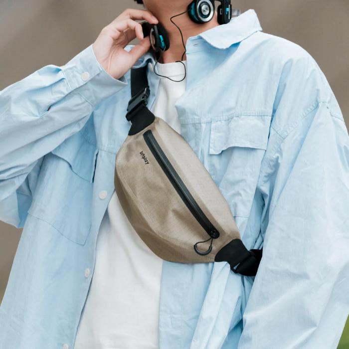 Person wearing a light blue shirt with a sand beige Bitplay AquaSeal IPX7 Waterproof Sling Bag with Strap Adapter and headphones, against a neutral background.
