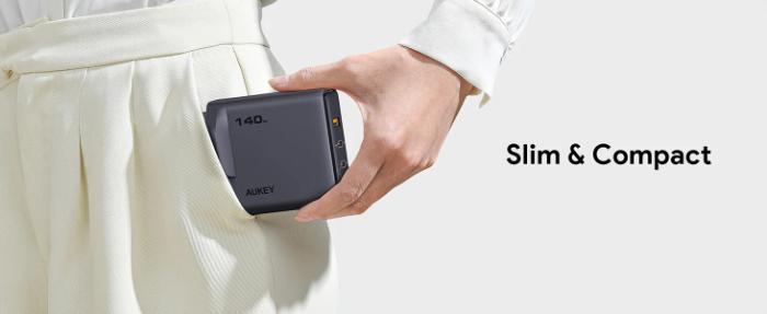 Hand holding a Gray Aukey PA-C6 Comet Mix 4 140W PD Wall Charger with GaN Technology with text 'Slim & Compact' on a white background