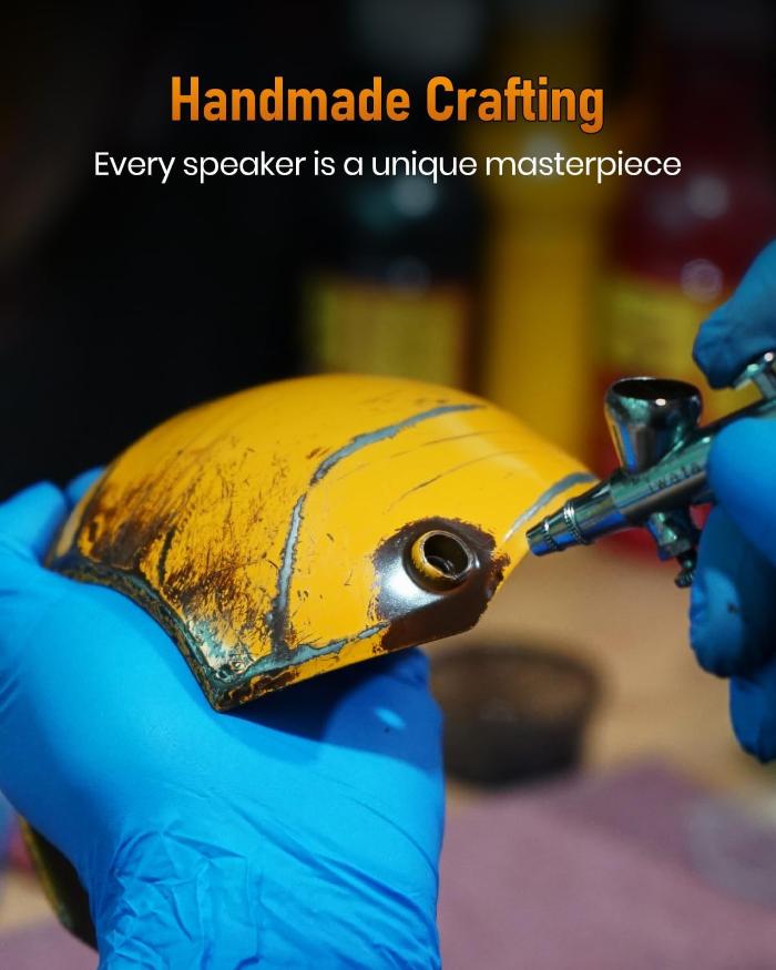 Person wearing blue gloves applying paint to a yellow GravaStar G1 Pro Mars Pro Wireless Bluetooth 5.0 Speaker (War Damaged Edition) with text 'Handmade Crafting' and 'Every speaker is a unique masterpiece'.